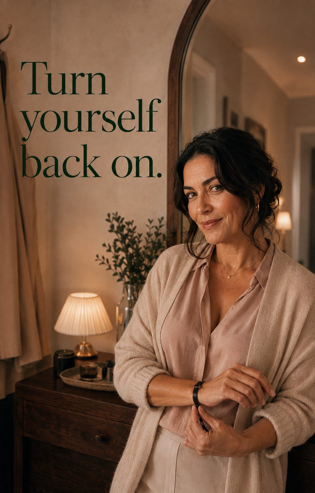 Turn yourself back on. A smiling woman in her fifties in a blush blouse and oatmeal cardigan, standing by a mirror in a warmly lit room.